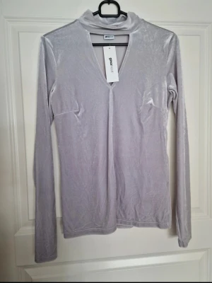 Ny ljuslila/grå sammets polotopp Gina Tricot med etikett - Supersnygg ljuslila polotopp från Gina Tricot i mjuk och glansig sammet. Toppen har lång ärm, lätt v-ringning och en smal passform som sitter snyggt på kroppen. Perfekt att styla med jeans eller kjol för en trendig look.