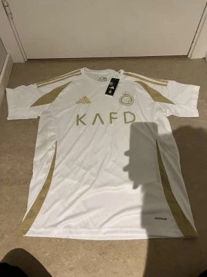 Vit och guld fotbollströja Adidas - Snygg vit fotbollströja från Adidas med gulddetaljer och klubbmärke på bröstet. Tröjan har korta ärmar, rund hals och guldiga ränder på axlarna. Tillverkad i lätt och ventilerande material, perfekt för träning eller match.
