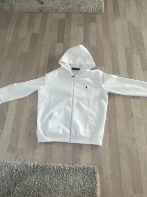 Vit hoodie från Polo Ralph Lauren - Snygg vit hoodie från Polo Ralph Lauren med dragkedja framtill och klassisk huva. Tröjan har två fickor på magen och den ikoniska lilla loggan broderad på bröstet. Perfekt för en clean och stilren look. Tillverkad i mjukt material för extra komfort.