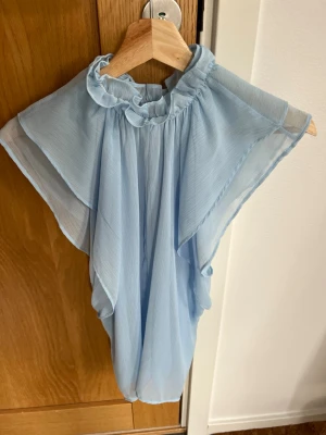 Ljusblå volangblus med rysch, från Zara - Supersöt ljusblå blus i ett tunt, transparent tyg med volangdetaljer och rysch vid halsen, från Zara. Blusen har en luftig och flowy passform och är ärmlös, perfekt för en trendig och fräsch look. Materialet känns lätt och svalt mot huden. Blusen är endast använd en gång, i princip som ny🩵!  
