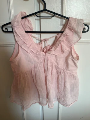 Ljusrosa volangtop från Gina Tricot - Supersöt ljusrosa ärmlös topp från Gina Tricot med volangkant runt urringningen och knytning i ryggen. Tillverkad i ett tunt, luftigt tyg som känns perfekt för sommaren. Passar dig som gillar romantisk och feminin stil.