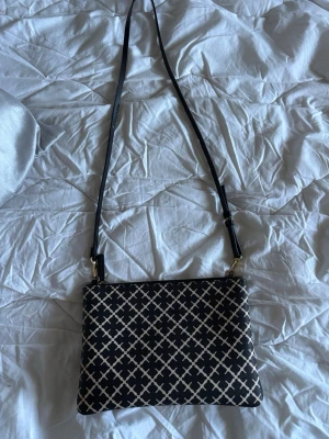 Svart axelväska med vitt mönster - Snygg svart axelväska med vitt grafiskt rutmönster och justerbar axelrem. Väskan har guldfärgade detaljer och en rektangulär form. Perfekt för dig som gillar stilrena accessoarer med en twist.