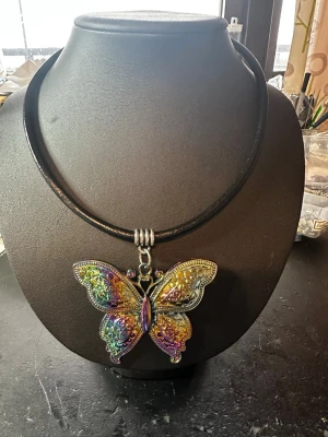 Regnbågsskimrande fjärilshalsband - Unikt halsband med en stor fjärilsberlock i metall som skimrar i regnbågens färger. Hängs på ett svart läderrem som ger en cool kontrast till den detaljerade fjärilen. Perfekt för dig som vill sticka ut med ett färgstarkt och fantasifullt smycke.