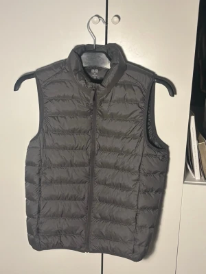 Grå dunväst från Uniqlo - Snygg grå dunväst från Uniqlo med quiltad design och hög krage. Västen har dragkedja framtill och är lätt vadderad, perfekt för lager-på-lager. Enkel och stilren look som funkar till det mesta.