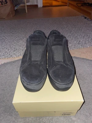 Svarta Axel arigato laceless skor - Stilrena svarta sneakers från Axel Arigato i mocka med diskret guldfärgad logga på sidan. Slip-on modell utan snörning och med svart sula. Perfekta för dig som gillar minimalistisk och clean stil.