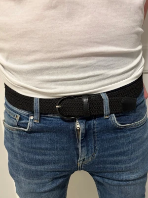 Svart flätat bälte i skinn och syntet - Snyggt svart flätat bälte med detaljer i skinn och metallspänne. Passar perfekt till jeans och ger en clean look. Bältet har en klassisk design och är lätt att justera för bästa passform. Hör av dig vid minsta intresse!💫⭐️