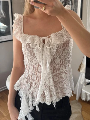 Vit spetsblus med volang och snörning - Romantisk vit topp i spets med volangkant och snörning framtill. Kan bäras både offshoulder och med axelband. Luftig och lätt, perfekt för sommarens outfits. Asymmetrisk nederkant ger en unik touch.