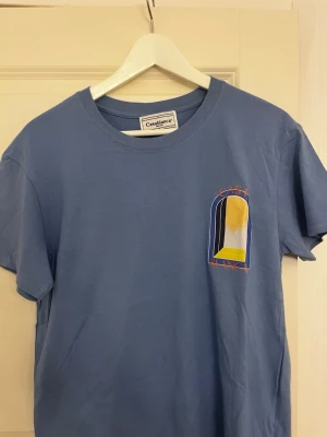 Blå t-shirt från Casablanca - Snygg blå t-shirt från Casablanca med färgglad grafisk print fram och bak i gult, vitt, blått och svart. T-shirten har rund hals och korta ärmar. Perfekt för dig som gillar streetwear och vill sticka ut med cool design.