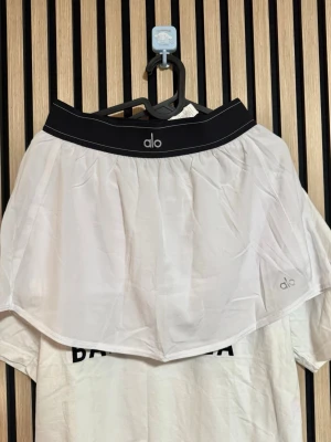 Vit kortkjol från Alo med svart linning - Säljer en snygg vit kortkjol från Alo med bred svart elastisk linning och logga framtill. Kjolen är tillverkad i lätt och luftig polyester, perfekt för sportiga aktiviteter. Enkel och stilren design som är lätt att matcha.