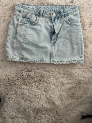 Ljusblå jeanskjol i kort modell - Säljer en ljusblå kort jeanskjol med klassiska fickor fram och bak, bälteshällor och knappstängning. Perfekt till sommaren och enkel att matcha med olika toppar. Snygg och tidlös denimstil!