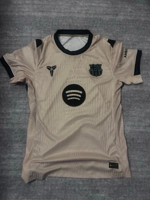 Beige FC Barcelona matchtröja Nike - Snygg beige FC Barcelona matchtröja från Nike med svarta detaljer och klubbmärke på bröstet. Tröjan har Spotify-logga framtill och UNHCR-tryck på ryggen. Korta ärmar och sportig passform, perfekt för fotboll eller träning.