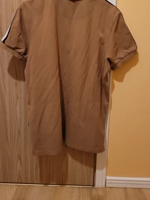 Beige pikétröja från BOSS - Snygg beige pikétröja från BOSS med korta ärmar och vit-svarta ränder längs axlarna. Tröjan har klassisk krage och knappar framtill. Perfekt för en stilren och avslappnad look.