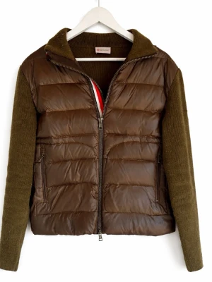 Moncler cardigan med stickade ärmar - Säljer en grön pufferjacka från Moncler med olivgröna stickade ärmar och krage. Jackan har dragkedja framtill, två fickor med dragkedja och Moncler-logga på ärmen. Perfekt för dig som vill ha en unik och snygg jacka med både quiltad och stickad känsla.