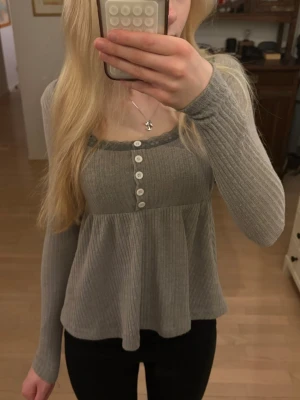 Grå ribbad långärmad tröja med spetsdetalj och knappar - Säljer en grå ribbad blus med långärmad design och vita knappar framtill. Blusen har en söt spetskant runt den fyrkantiga halsringningen och ett lätt utsvängt fall. Perfekt för dig som gillar en feminin och chill stil.