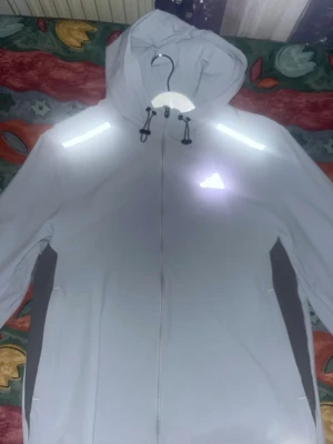 Ljusgrå vindjacka från Adidas - Snygg ljusgrå vindjacka från Adidas med reflekterande detaljer på axlar och bröst. Jackan har huva med dragsko, hel dragkedja framtill och svarta paneler på sidorna. Perfekt för blåsiga dagar och sportiga outfits.