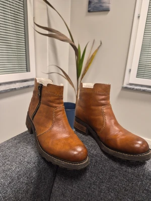 Rieker lättfodrade Kängor Brun Storlek 37 - Snygga bruna stövletter i läder med grov sula och låg klack. De har en praktisk dragkedja på sidan och är fodrade med mjukt, vitt foder för extra värme. Perfekta för kyligare dagar och ger en cool touch till din outfit.