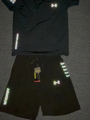 Svart träningsset från Under Armour - Snyggt svart träningsset från Under Armour med t-shirt och shorts. Båda plaggen har reflekterande loggor och text i regnbågsskimrande färg. Shortsen har dragsko i midjan och sidofickor. Perfekt för gymmet eller löprundan.