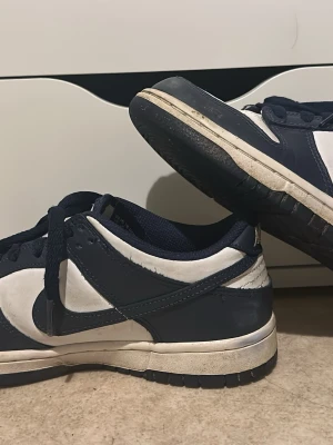 Nike Dunk Low blåvita sneakers - Säljer ett par Nike Dunk Low sneakers i blått och vitt. Skorna har klassisk låg siluett, vita partier i läder med mörkblå overlays och matchande blå snören. Ikonisk Nike Swoosh på sidan och perforerad tå för extra ventilation. Perfekta för en streetwear-look. Eur 38