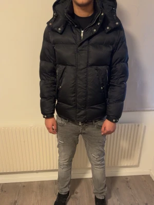 Svart quiltad pufferjacka från BOSS - Säljer en svart pufferjacka från BOSS med diskret mönster och quiltad design. Jackan har huva, dragkedja framtill och två stora fickor med knappstängning. Perfekt för kalla dagar och riktigt snygg till streetwear-stilen.