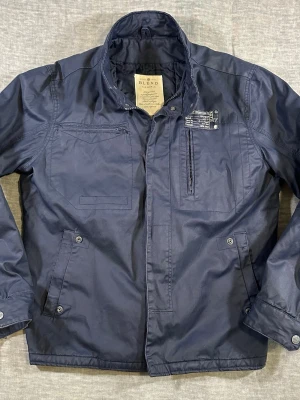 Utility Field Jacket XL Navy Workwear Military Jacket Blend - Blend utility field jacket in navy blue, size XL. Water-resistant cotton shell with a clean military-inspired design. Features multiple utility pockets, chest zip pocket, and snap + zip front closure. Lightweight but durable – perfect for everyday wear, layering, or that rugged workwear look.  Measurements (cm, laid flat): S2S 51 / P2P 62 / Waist 59 / Sleeve 68 / Length 74  Good used condition with normal wear.