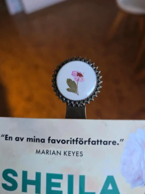 Metallbokmärke med blomdekor - Snyggt bokmärke i metall med en rosa blomma med gröna blad. Perfekt för dig som vill ha något unikt och stilrent till dina böcker. Bokmärket har en antik mässingsfärg och en dekorativ kant runt motivet.