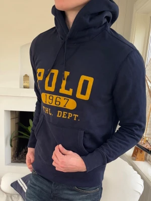 Mörkblå hoodie från Polo Ralph Lauren - Mörkblå hoodie med stor gul POLO 1967 ATHL. DEPT. print på bröstet. Klassisk känguruficka framtill, dragsko i huvan och ribbade muddar. Perfekt för en avslappnad och sportig stil.