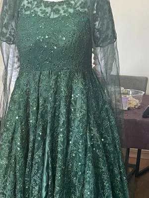 Mörkgrön långklänning med spetsdetaljer - Elegant mörkgrön långklänning med broderade spetsdetaljer och pärlor. Klänningen har korta ärmar i transparent tyg och en vacker, utsvängd kjol. Perfekt för bal eller festliga tillfällen. Materialet är troligtvis polyester eller syntet med dekorativa inslag.