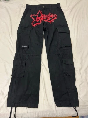 Svarta cargobyxor med röd patch - Säljer ett par svarta cargobyxor med stor röd haj-patch och text på baksidan. Byxorna har flera stora fickor på sidorna och dragsko vid bensluten. Perfekta för dig som gillar streetwear och vill sticka ut med en unik detalj.