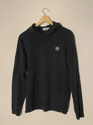 Svart hoodie från Stone Island - Snygg svart hoodie från Stone Island med klassisk patch-logga på bröstet. Hoodien har en enkel och stilren design med huva och långa ärmar. Tillverkad i mjukt bomullsmaterial som är skönt att bära. Perfekt för dig som gillar streetwear och vill ha en clean look.