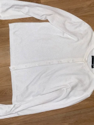 Vit basic kofta med knappar - En enkel och stilren vit kofta med rund hals och knäppning framtill. Långärmad modell i mjukt, tunt stickat material. Perfekt att slänga över en topp eller klänning för en clean look.