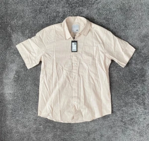 Beige kortärmad skjorta från H&M - Snygg beige skjorta från H&M med korta ärmar och klassisk krage. Skjortan har knappar framtill och en bröstficka. Perfekt för en avslappnad och stilren look.