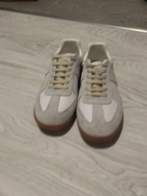 Vita och beige sneakers med gummisula - Snygga sneakers i vitt och beige med klassisk retrostil. Ovandelen är i mocka och textil, och skorna har en brun gummisula med mönstrad undersida för bra grepp. Perfekta för dig som gillar stilrena och tidlösa sneakers.