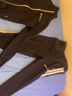 Palm Angels svart dress båda är ÄKTA och i strl S - Säljer ett svart träningsset från Palm Angels med vita detaljer och dragkedja på jackan. Setet består av en zip-jacka och matchande byxor med vita ränder längs sidorna och Palm Angels-logga på både jacka och byxa. Perfekt för sport eller chill. Materialet är mjukt och stretchigt.