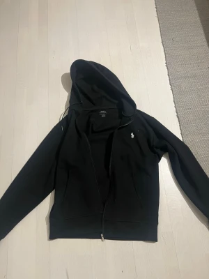 Polo Ralph Lauren Svart hoodie  - Snygg svart hoodie från Polo Ralph Lauren med dragkedja och klassisk vit logga på bröstet. Tillverkad i mjukt bomullsmaterial och har två fickor framtill samt en stor huva. Perfekt för en avslappnad och stilren look.