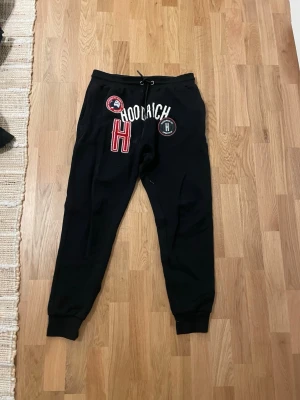 Svarta mjukisbyxor från Hoodrich - Svarta mjukisbyxor från Hoodrich med broderade patches och tryck i vitt, rött och svart. Byxorna har resår och dragsko i midjan samt muddar vid bensluten. Perfekta för en avslappnad och streetinspirerad stil.