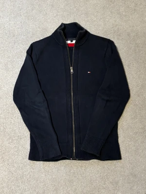 Fullzip Tommy Hilfiger - Snygg marinblå ziptröja från Tommy Hilfiger med hög krage och diskret logga på bröstet. Tröjan är ribbstickad med långa ärmar och har breda muddar vid ärmslut och nederkant. Perfekt för en stilren och avslappnad look.