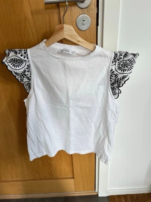 Vit topp med broderade volangärmar MNG - Säljer en vit topp från Mango, med unika volangärmar som har svart broderat mönster. Toppen är ärmlös med breda axelvolanger och har en rund halsringning. Perfekt för dig som vill ha något extra till din outfit. Endast använd ett fåtal gånger, som ny💖! 