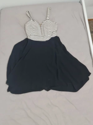 Randig kortklänning med dragkedja - Snygg kortklänning med svart kjol och vit topp med svarta ränder. Klänningen har smala axelband och en dekorativ dragkedja framtill. Perfekt för dig som gillar en enkel men trendig stil.