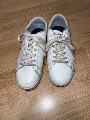 Vita sneakers från Lacoste - Säljer ett par klassiska vita sneakers från Lacoste med låga skaft och vita snören. Skorna har den ikoniska krokodilloggan på sidan och är tillverkade i slätt läder med diskreta sömmar. Perfekta för en clean och stilren look. Snören kan bytas använd bara en sommar.