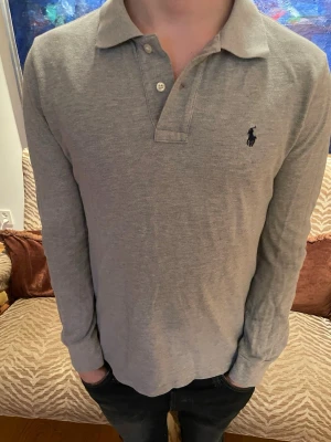 Grå långärmad pikéskjorta Polo Ralph Lauren - Snygg grå långärmad pikéskjorta från Polo Ralph Lauren med klassisk krage och knappar framtill. Ikoniska loggan broderad på bröstet. Tillverkad i mjuk bomull som känns skön mot huden. Perfekt för en clean och stilren look. Pris är diskuterbart!🙌