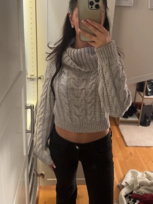 Grå stickad polo tröja - En snygg grå croppad stickad tröja med bred polokrage och kabelstickat mönster. Tröjan har långa ärmar och ribbade muddar. Perfekt för att skapa en trendig look under kyliga dagar.( har använt 1 gång)