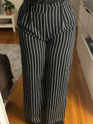 Svarta vida leggings med vita ränder - Säljer ett par svarta leggings med vita vertikala ränder och hög midja. Modellen är lös och har breda ben, vilket ger en avslappnad och trendig look. Perfekta för dig som gillar streetstyle och vill ha något bekvämt men ändå snyggt.