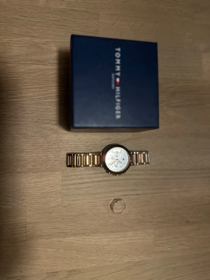 Guldfärgad klocka Tommy Hilfiger - Stilren guldfärgad klocka från Tommy Hilfiger med metallarmband och rund urtavla i silver. Klockan har flera små urtavlor och klassisk design. Perfekt accessoar för dig som vill ha något snyggt och tidlöst på handleden. Och en silver ring som sitter fint på en i topp skick 