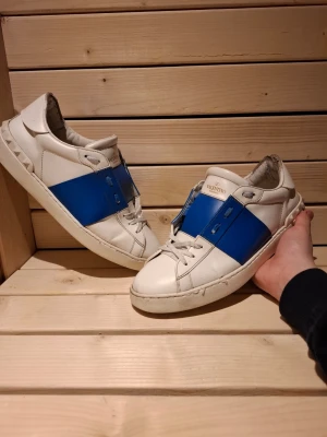 Valentino Open vita sneakers med blå detalj i storlek 43 - Snygga vit blåa Valentino Opens från Valentino Garavani med en bred blå rem över mitten. Skorna har klassisk låg profil, vita snören och diskret logga på plösen. Tillverkade i läder med stilren design som sticker ut med den blåa detaljen. De är i riktigt bra skick och väl omhändetagna, de är i storlek 43. Bara att fråga om ni vill ha mer bilder eller diskutera priset, Skickar samt svarar väldigt fort 😀