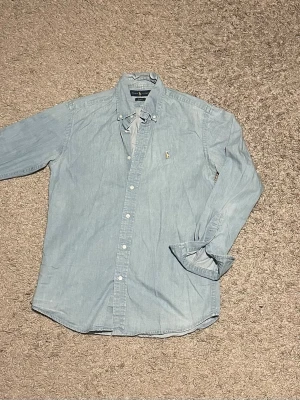 Ljusblå skjorta från Ralph Lauren - Säljer en ljusblå slim fit skjorta från Ralph Lauren med klassisk button-down krage och broderad logga på bröstet. Skjortan är tillverkad i mjukt bomullstyg och har långa ärmar med manschetter. Perfekt för en clean och stilren look.