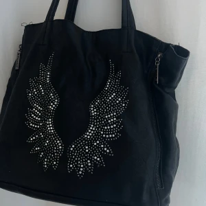 ANGEL VÄSKA  - Super snygg svart väska med änglavingar på, från ETERNAL. Använd men i gott skick! Går att ha som axelremsväska men medföljer även långt band. Necessär inuti som går att ta ut. Perfekt som skol eller jobb väska då den är lite större!