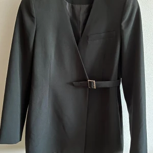 Kavaj med silverdetalj  - Svart kavaj med en silverdetalj/knäppe. Knäppet har inte så stor funktion men kavajen har en inre knapp som man använder. Finns inget märke eller storlek på kavajen men jag skulle säga att den passar storlek 36-38. 