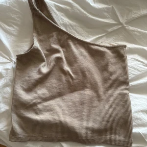 Beige ribbad oneshoulder topp från Gina Tricot - En stilren beige ribbad oneshoulder topp från Gina Tricot. Toppen har en asymmetrisk design med en axelbandslös sida och är tillverkad i ett mjukt och stretchigt material.
