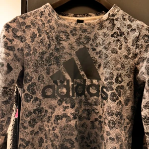 Grå leopardmönstrad tröja från Adidas - En grå leopardmönstrad tröja från Adidas med svart logotyp på framsidan. Tröjan har långa ärmar och en rund halsringning.