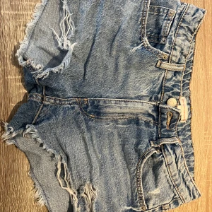 Blå jeansshorts från Gina Tricot - Ett par blå jeansshorts från Gina Tricot med slitna detaljer och fransiga kanter. Shortsen har en knapp och dragkedja framtill samt fem fickor.